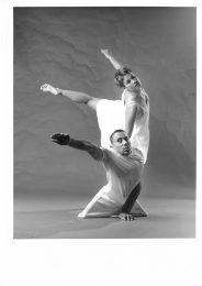 2 dancers are close to each other, side on to the camera. The dancer behind has her hand on the other's shoulder with her leg lifted behind and her left arm rised. The male dancer in front has his right arm raised towards the camera.