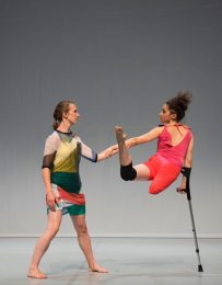 Dancer wearing bright pink leans on crutch as her back leg lefts high, she reaches back to dancer standing in multicoloured top