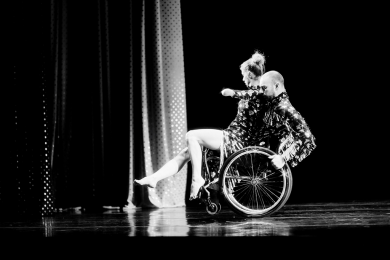 A female dancer sits on the lap of a male in a wheelchair, both dressed for a show.