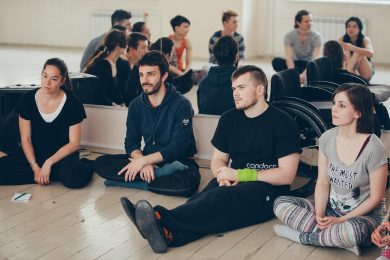 4 people sit against mirror listening, we see the reflection of other people in the mirror