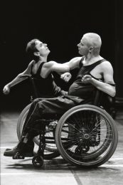 2 dancers are facing each other in wheelchairs. Their arms are linked. Picture is black and white.