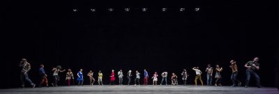 A long row of people stand across the black stage. Each person wears brightly coloured clothing.