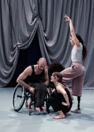 3 people in front of a grey draped curtain which is hoisted up in the centre. Liv is in the front and is crouching down, Joel is seated in a wheelchair and leaning forwards towards Liv. Mickaella is standing to the side with her front knee lifted and arms long above her ahead.