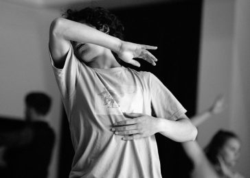 Black and white photo. A young dancer leans back, slightly twisting at the torso. His left hand presses gently against his ribs, and his face is hidden behind his right elbow which is pointing upward.