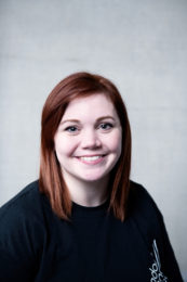 Hannah Raynor. A headshot of a smiling person with long reddish brown hair who is wearing a black t-shirt